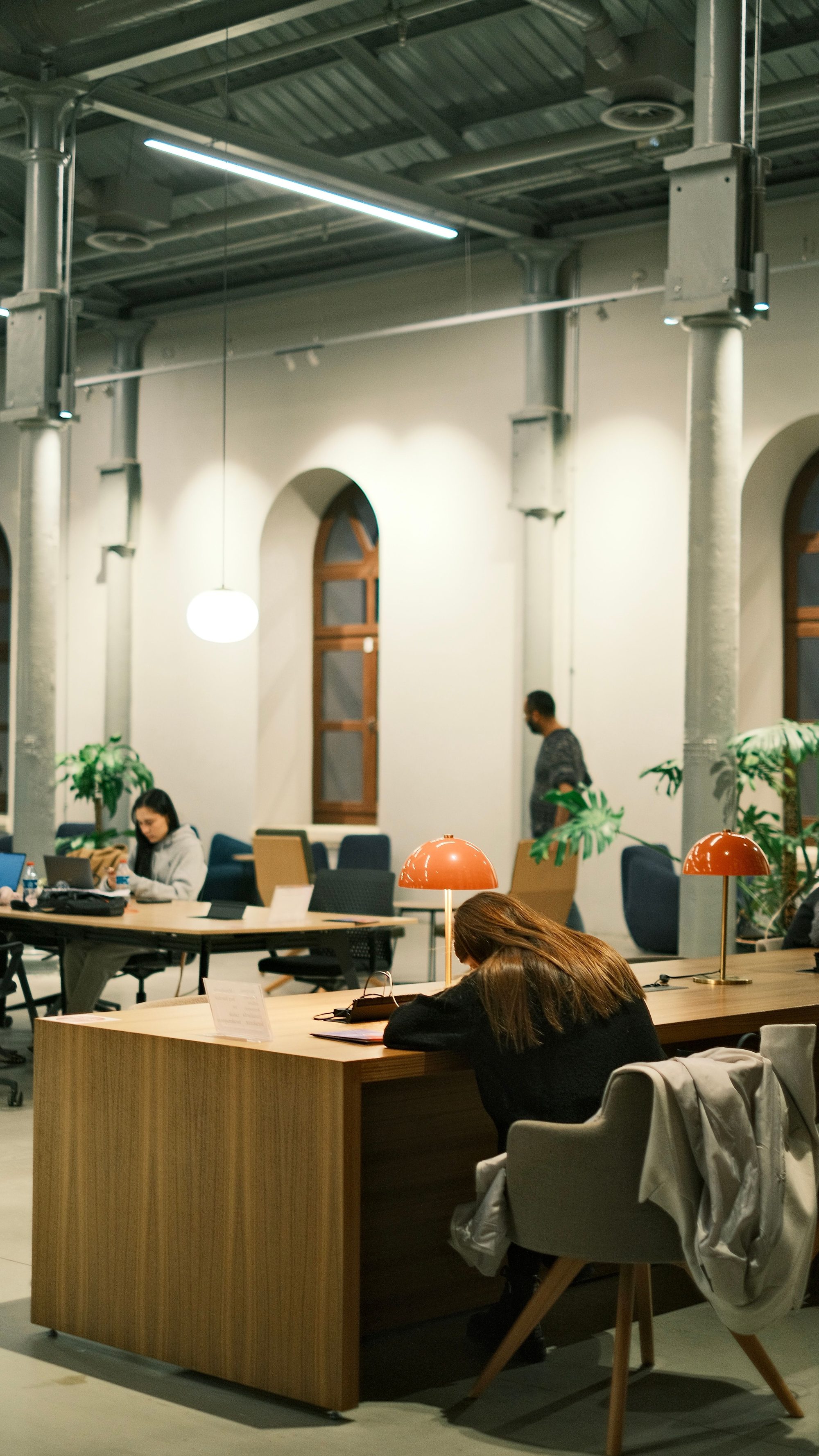Members working at long wooden desks beneath original arched windows and industrial pendant lighting inside the Northline Collective loft, with warm coral desk lamps and potted plants throughout the open workspace.