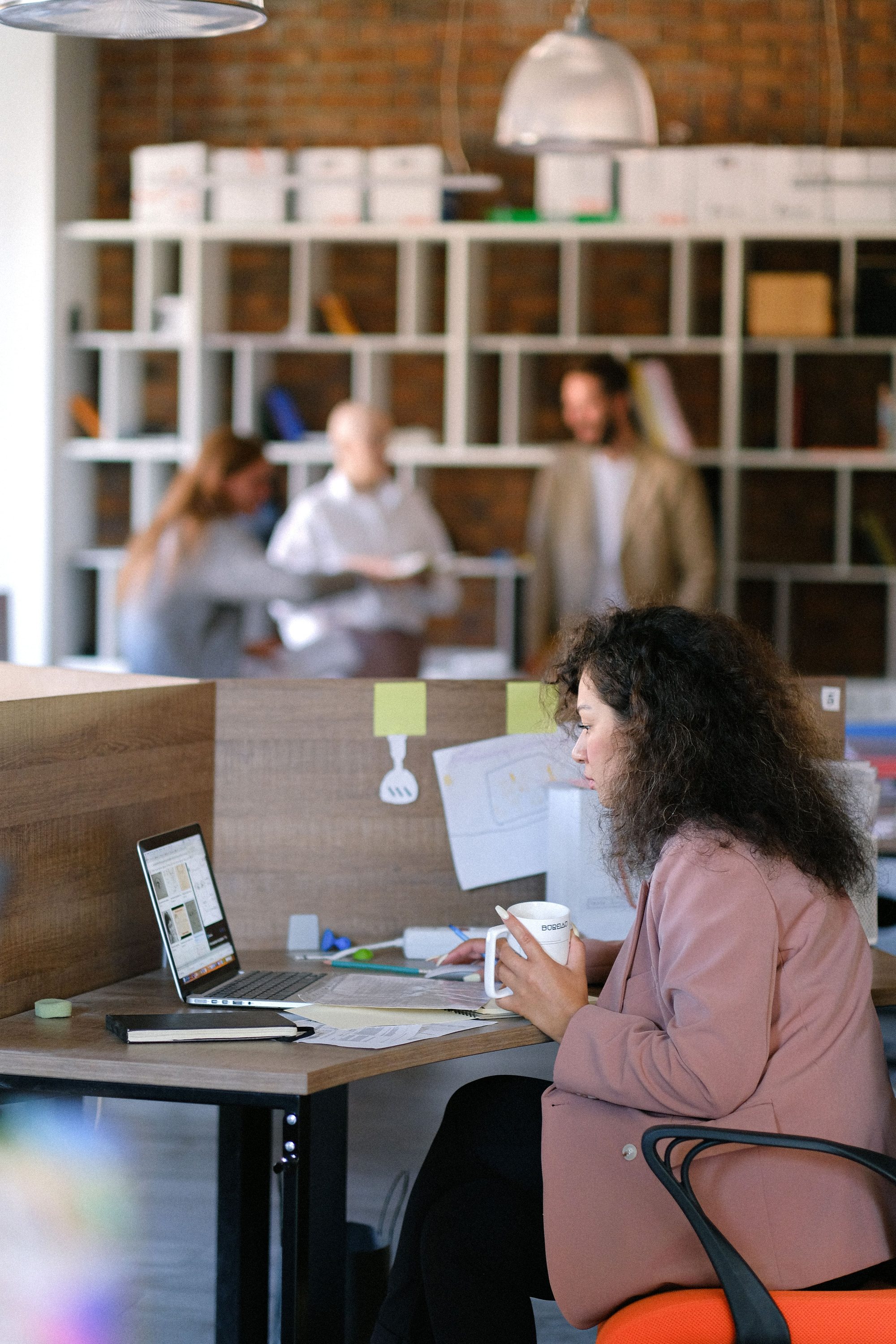 A Northline Collective member reviewing notes over coffee at a window-side desk, with teammates catching up near the shared resource shelving in the background.